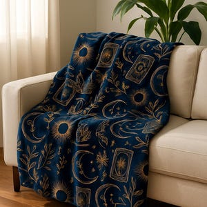 May include: A deep navy blue throw blanket featuring a repeating gold-toned pattern of suns, crescent moons, rectangular tarot-style cards, and botanical leaf sprigs. The fabric has a soft, plush texture and is draped over a light-colored sofa.
