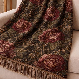 May include: A woven throw blanket featuring a dark background with a repeating pattern of deep red roses and green foliage. The fabric has a textured appearance and is finished with long, light brown fringe along the bottom edge of the textile.