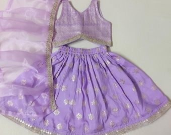 Op de afbeelding: Een lavendelkleurige tweedelige outfit bestaande uit een mouwloze crop top en een geplooide rok, beide met een metallic goudkleurig bloemenpatroon. De set bevat een bijpassende transparante lavendelkleurige sjaal met een gouden rand.