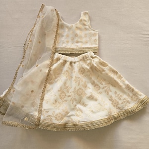 Puede incluir: Conjunto de tres piezas de color crema, que incluye un top corto, una falda y un velo transparente. El conjunto está adornado con un estampado floral dorado y ribetes dorados. La falda es acampanada y el top tiene cuello redondo. El velo es transparente.