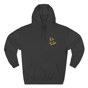 Puede incluir: Sudadera con capucha gris oscuro con bolsillo delantero y capucha. El texto "Bee A Light" está bordado en amarillo en la parte superior izquierda del pecho. La sudadera está hecha de un material suave y cómodo.