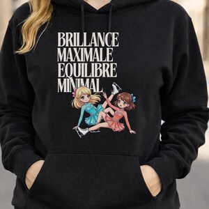Puede incluir: Sudadera con capucha negra con un gráfico de dos patinadoras artísticas de dibujos animados con vestidos azules y coral. El texto "BRILLANCE MAXIMALE EQUILIBRE MINIMAL" está impreso en blanco sobre las patinadoras. La sudadera tiene un bolsillo delantero y una capucha con cordón.