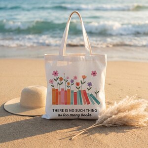 Op de afbeelding: Een crèmekleurige canvas draagtas met een kleurrijke afbeelding van boeken waar bloemen uit groeien. De tekst op de tas luidt: "THERE IS NO SUCH THING as too many books." De tas is voorzien van twee lange schouderbanden en een platte bodem.