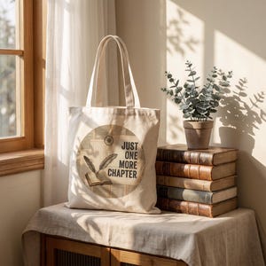 May include: A cream-colored canvas tote bag featuring a circular graphic with a vintage book, a quill pen, and the text "JUST ONE MORE CHAPTER" printed in black. The bag has two long shoulder straps and is constructed from a durable fabric material.