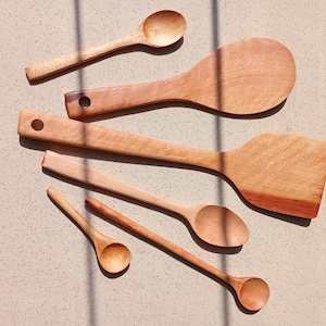 May include: A selection of wooden kitchen utensils, including spoons and spatulas, displayed on a light surface. The utensils are of various sizes and shapes, with a natural wood grain. The image is lit with natural light, creating shadows.