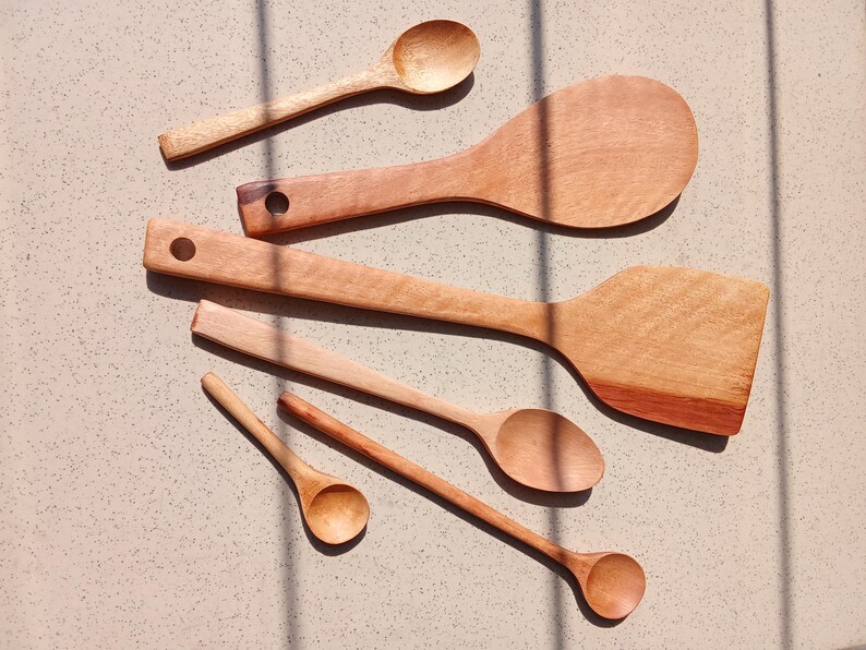 May include: A selection of wooden kitchen utensils, including spoons and spatulas, displayed on a light surface. The utensils are of various sizes and shapes, with a natural wood grain. The image is lit with natural light, creating shadows.