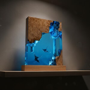 May include: A rectangular wooden and resin art piece featuring a marine scene with manta rays. The artwork has a wooden frame with a natural wood grain. The interior is a vibrant blue, with a column and manta rays.