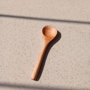 May include: A wooden spoon with a rounded bowl and a long, slender handle. The spoon is made of light-colored wood and is set against a speckled, neutral-toned surface. The spoon is angled, casting a shadow.