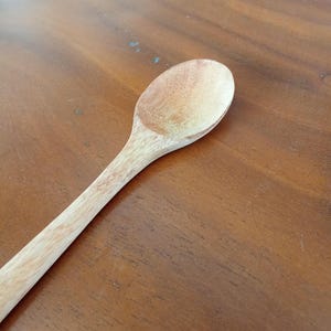May include: A wooden spoon with a long handle and a rounded bowl. The spoon is made of light-colored wood and has a smooth, natural finish. The spoon is designed for cooking and serving food.