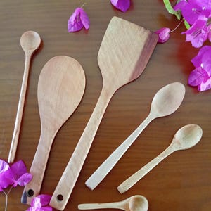 May include: A selection of wooden kitchen utensils, including spoons and a spatula, displayed on a brown surface. The utensils are of various sizes and shapes, with visible wood grain. Bright purple flowers are scattered around the utensils, adding colour.
