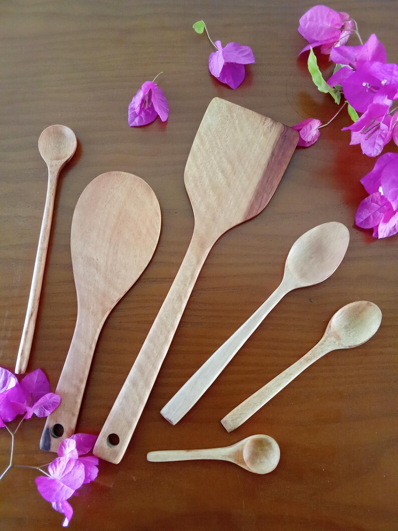 May include: A selection of wooden kitchen utensils, including spoons and a spatula, displayed on a brown surface. The utensils are of various sizes and shapes, with visible wood grain. Bright purple flowers are scattered around the utensils, adding colour.