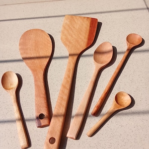May include: A selection of wooden kitchen utensils, including spoons and a spatula, displayed on a light surface. The items vary in size and shape, with visible wood grain and natural tones.