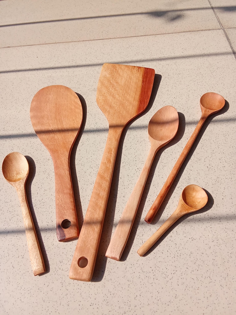 May include: A selection of wooden kitchen utensils, including spoons and a spatula, displayed on a light surface. The items vary in size and shape, with visible wood grain and natural tones.
