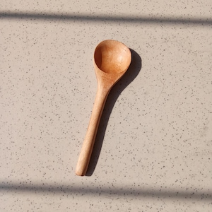May include: A wooden spoon with a rounded bowl and a long handle. The spoon is made of light-coloured wood and is positioned on a speckled surface. The spoon's design is simple and functional, suitable for kitchen use.