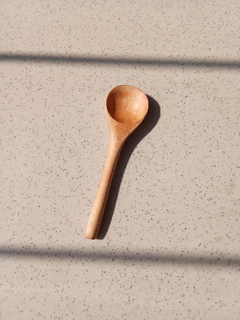 May include: A wooden spoon with a rounded bowl and a long handle. The spoon is made of light-coloured wood and is positioned on a speckled surface. The spoon's design is simple and functional, suitable for kitchen use.