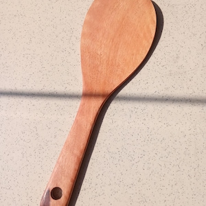 May include: A wooden rice paddle with a long handle and a rounded, flat head. The paddle is made of light brown wood and has a hole in the handle for hanging. The kitchen utensil is designed for serving rice.