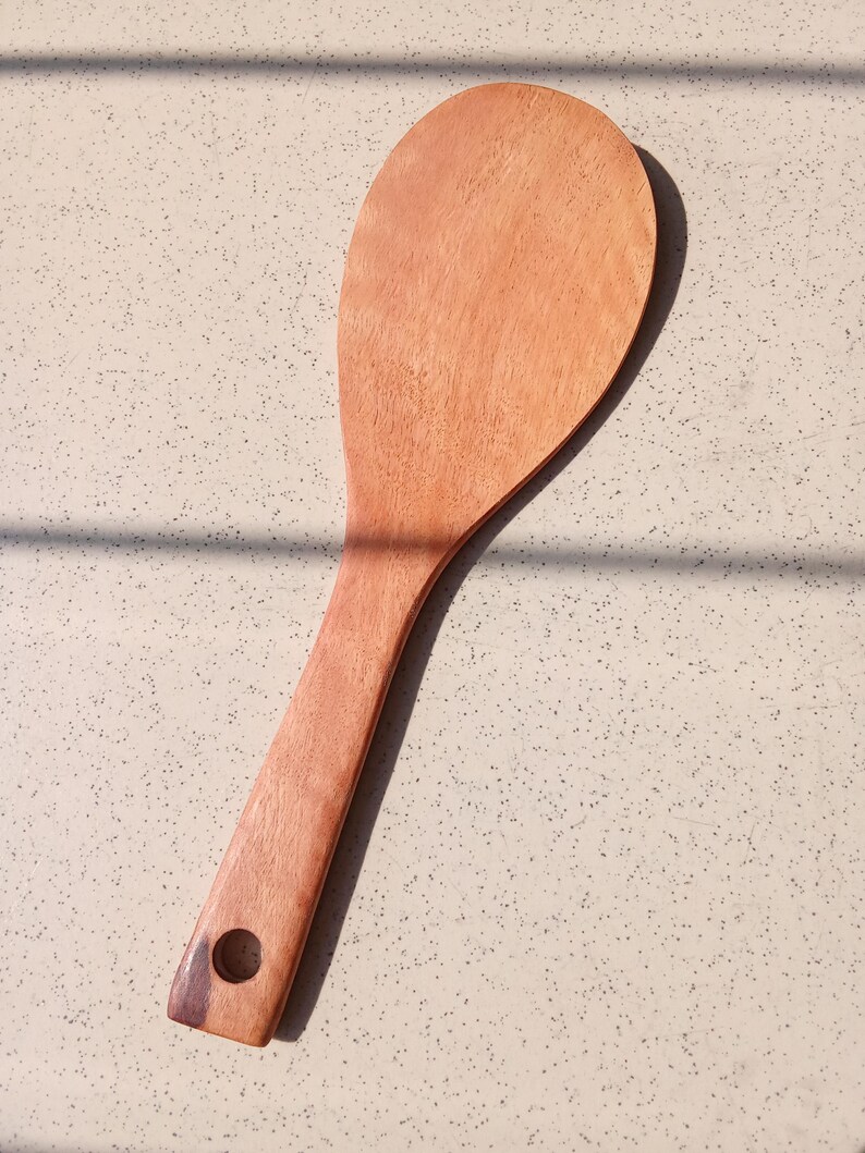 May include: A wooden rice paddle with a long handle and a rounded, flat head. The paddle is made of light brown wood and has a hole in the handle for hanging. The kitchen utensil is designed for serving rice.