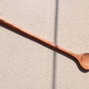 May include: A long wooden spoon with a rounded bowl and a slender handle. The spoon is made of light brown wood and is lying on a speckled, light-coloured surface. The spoon's shadow is visible.