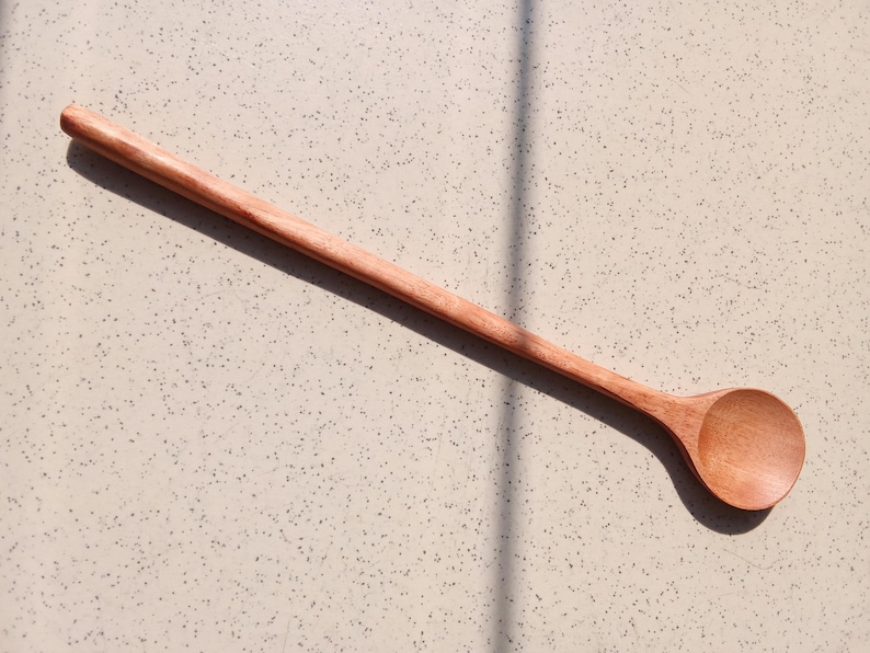 May include: A long wooden spoon with a rounded bowl and a slender handle. The spoon is made of light brown wood and is lying on a speckled, light-coloured surface. The spoon's shadow is visible.