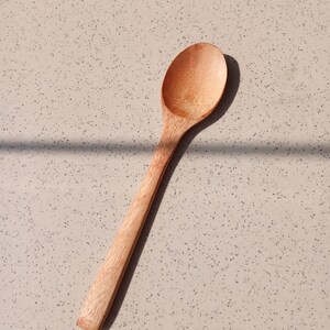 May include: A wooden spoon with a long handle and a rounded bowl. The spoon is made of light-coloured wood and is set against a speckled, neutral-toned surface. The spoon is angled diagonally across the frame.