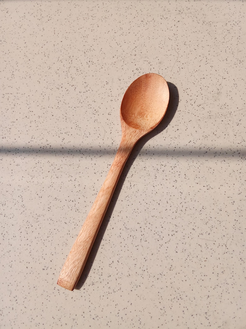 May include: A wooden spoon with a long handle and a rounded bowl. The spoon is made of light-coloured wood and is set against a speckled, neutral-toned surface. The spoon is angled diagonally across the frame.
