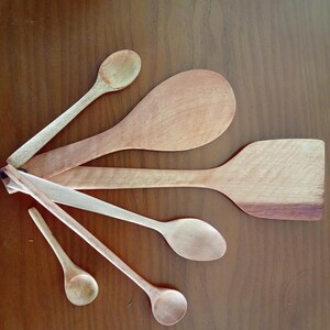 May include: A selection of wooden kitchen utensils, including spoons and a spatula, displayed on a brown surface. The spoons are of various sizes and shapes, in light brown hues. The spatula has a darker brown edge.