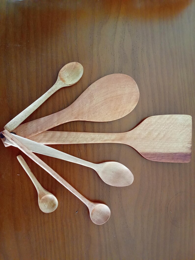 May include: A selection of wooden kitchen utensils, including spoons and a spatula, displayed on a brown surface. The spoons are of various sizes and shapes, in light brown hues. The spatula has a darker brown edge.