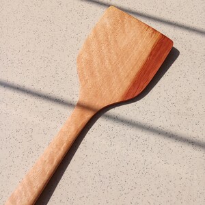 May include: A wooden spatula with a broad, flat head and a long, thin handle. The spatula is made of light brown wood with a reddish-brown edge, and it is on a speckled surface. The spatula is designed for cooking and food preparation.
