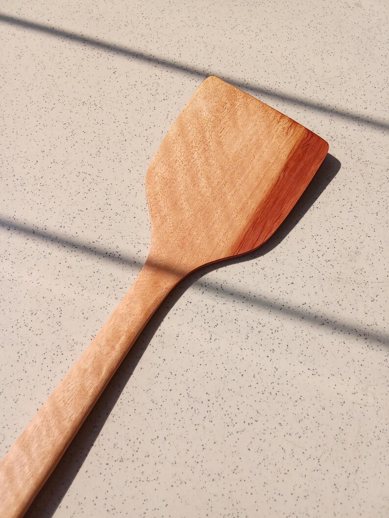 May include: A wooden spatula with a broad, flat head and a long, thin handle. The spatula is made of light brown wood with a reddish-brown edge, and it is on a speckled surface. The spatula is designed for cooking and food preparation.