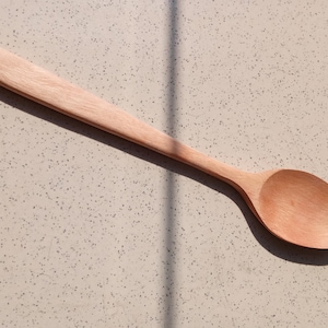 May include: A light brown wooden spoon with a long handle and a rounded bowl. The spoon is made of a light-coloured wood and is lying on a speckled surface. The spoon is designed for cooking and serving.