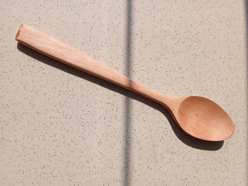 May include: A light brown wooden spoon with a long handle and a rounded bowl. The spoon is made of a light-coloured wood and is lying on a speckled surface. The spoon is designed for cooking and serving.