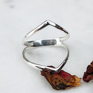 May include: A silver ring with a double V-shaped design. The ring is made of shiny metal and sits on a white surface with a few dried petals. The ring's design is simple and modern, suitable for everyday wear.