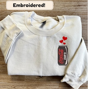 May include: A light gray sweatshirt with an embroidered Coca-Cola Zero can design and three red hearts. The sweatshirt has the word "Embroidered!" in a white box at the top. The design is on the front of the sweatshirt.