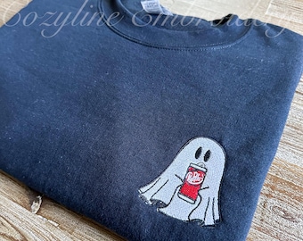 May include: Navy blue sweatshirt with an embroidered ghost holding a Dr. Pepper can. The ghost is white with black eyes and the soda can is red and white. The sweatshirt is folded on a wooden surface.