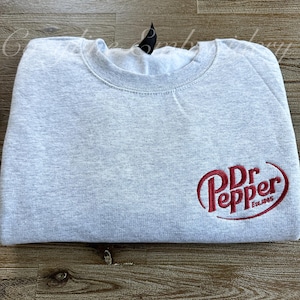 May include: A light gray sweatshirt with a crew neck. The sweatshirt features the Dr Pepper logo in red on the front, with the text "Est. 1885" below the logo. The sweatshirt is folded neatly.