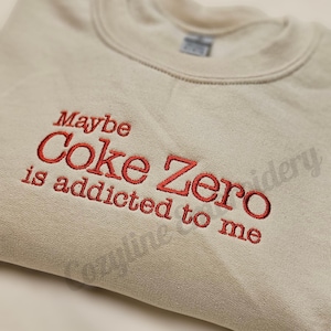 May include: A beige sweatshirt with the embroidered phrase "Maybe Coke Zero is addicted to me" in red thread. The crew neck sweatshirt is made of a soft material and is suitable for casual wear.