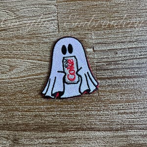 May include: Embroidered patch featuring a white ghost holding a Coca-Cola can. The ghost has black eyes and a red outline. The Coca-Cola can is red and white, with the Coca-Cola logo visible. The patch is on a wooden surface.