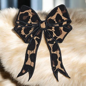 May include: A decorative bow with a leopard print pattern in shades of brown and black. The bow is made of fabric and has a detailed, embroidered design. The bow is likely used as an accessory.