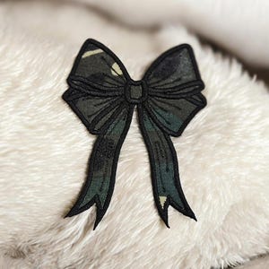May include: A black bow-shaped patch with a camouflage pattern in shades of green and brown. The patch has a black outline and long, flowing tails. The bow is centered on a fluffy, white surface.