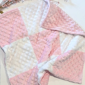 May include: A soft, plush blanket featuring a checkered pattern of alternating light pink and white squares. The fabric has a raised, dimpled texture across the entire surface, creating a tactile, bumpy appearance throughout the material.