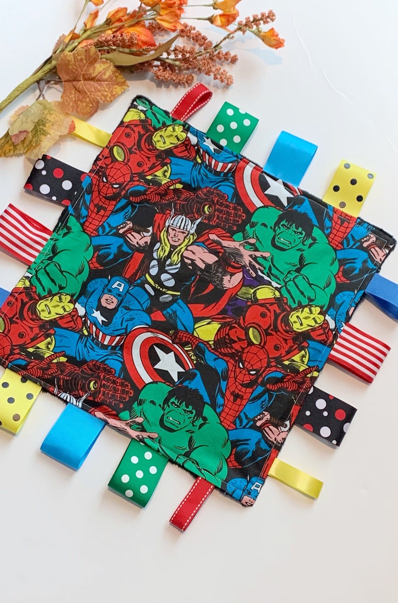 Op de afbeelding: Een zacht, vierkant babydekentje met een kleurrijke comic-print van superhelden zoals Spiderman, Hulk, Captain America en Thor. Het dekentje heeft meerdere kleurrijke satijnen linten aan de randen.