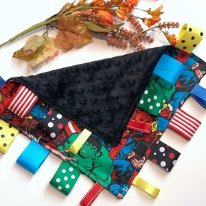 Op de afbeelding: Een zwart-wit babyzintuiglijk speelgoed met een superhelden thema. Het speelgoed heeft een zachte zwarte stof met een textuur en een verscheidenheid aan kleurrijke linten eraan bevestigd. De linten zijn gemaakt van verschillende materialen, waaronder satijn, stippenstof en gestreepte stof. Het speelgoed is ontworpen om de zintuigen van een baby te stimuleren en exploratie aan te moedigen.