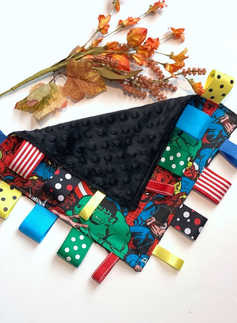 Op de afbeelding: Een zwart-wit babyzintuiglijk speelgoed met een superhelden thema. Het speelgoed heeft een zachte zwarte stof met een textuur en een verscheidenheid aan kleurrijke linten eraan bevestigd. De linten zijn gemaakt van verschillende materialen, waaronder satijn, stippenstof en gestreepte stof. Het speelgoed is ontworpen om de zintuigen van een baby te stimuleren en exploratie aan te moedigen.