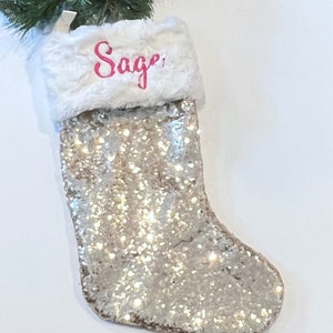 May include: A Christmas stocking with a silver glitter design and a white faux fur cuff. The name "Sage" is written in pink script across the cuff. Green and gold Christmas ornaments and greenery are visible in the background.