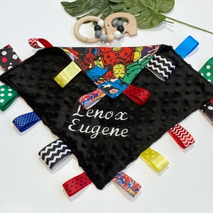 Może przedstawiać: Czarno-biały kocyk bezpieczeństwa dla niemowląt z nadrukiem superbohater&oacute;w i kolorowymi wstążkami. Kocyk jest spersonalizowany imieniem "Lenox Eugene".