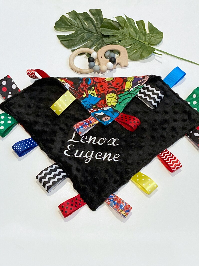 Może przedstawiać: Czarno-biały kocyk bezpieczeństwa dla niemowląt z nadrukiem superbohater&oacute;w i kolorowymi wstążkami. Kocyk jest spersonalizowany imieniem "Lenox Eugene".
