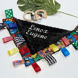 Może przedstawiać: Czarno-biały kocyk bezpieczeństwa dla niemowląt z motywem superbohater&oacute;w, z czarnym pluszowym środkiem. Kocyk ma r&oacute;żne kolorowe wstążki przymocowane do krawędzi i imię "Lenox Eugene" wyszyte na środku białym kolorem.