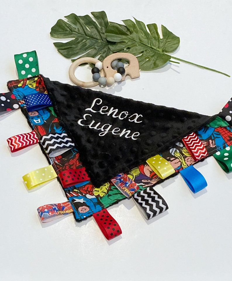 Może przedstawiać: Czarno-biały kocyk bezpieczeństwa dla niemowląt z motywem superbohater&oacute;w, z czarnym pluszowym środkiem. Kocyk ma r&oacute;żne kolorowe wstążki przymocowane do krawędzi i imię "Lenox Eugene" wyszyte na środku białym kolorem.