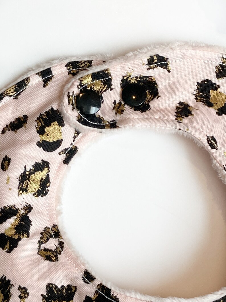 May include: Pink baby bib with a gold and black leopard print and two black snap closures.