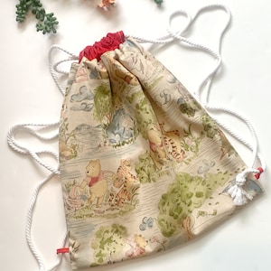 May include: A drawstring backpack featuring a beige fabric with a repeating illustration of Winnie the Pooh characters in a forest setting. The bag has a red fabric top opening and white braided rope straps with knotted ends at the bottom corners.